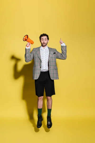 Hombre de moda señalando con el dedo y sosteniendo el altavoz mientras salta sobre fondo amarillo - foto de stock