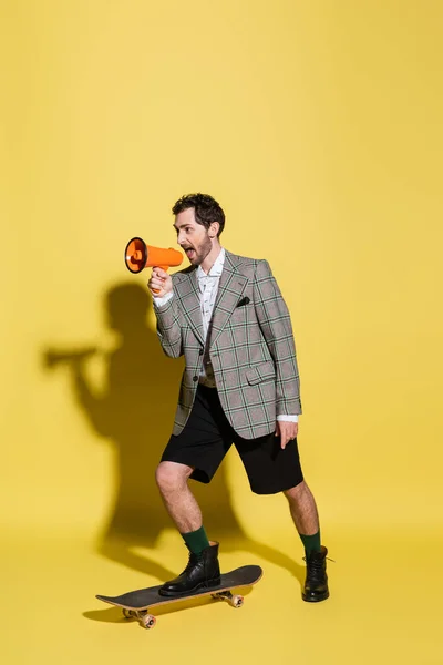 Trendy man holding loudspeaker and skateboarding on yellow background — Stock Photo