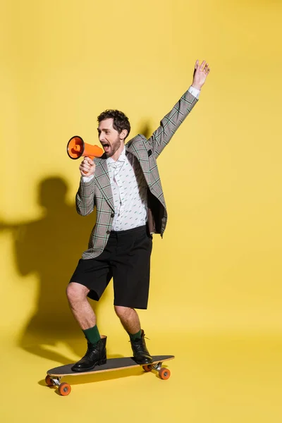 Fashionable man holding loudspeaker while skateboarding on yellow background — Stock Photo