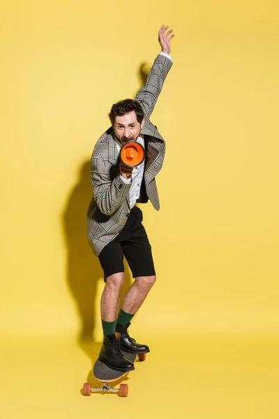 Stylish man holding loudspeaker and riding skateboard on yellow background — Stock Photo