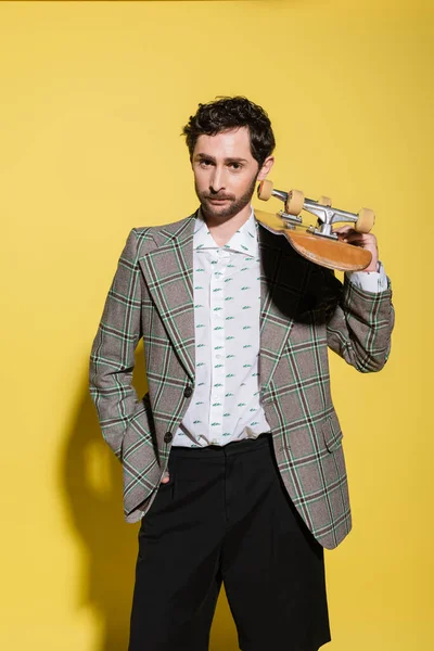 El hombre de moda en la chaqueta que sostiene el monopatín y mirando a la cámara en el fondo amarillo - foto de stock