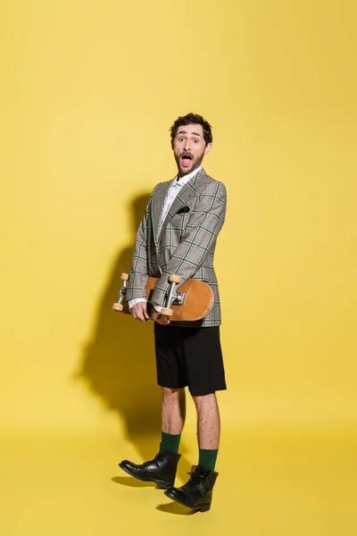 Emocionado e elegante homem de casaco e shorts segurando skate no fundo amarelo — Fotografia de Stock