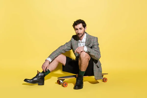 Charming guy in checkered jacket sitting on skateboard on yellow background — Stock Photo