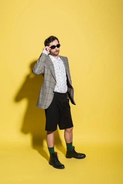 Fashionable man in shorts and jacket holding sunglasses on yellow background — Stock Photo