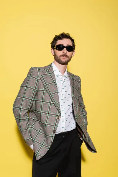 Trendy model in sunglasses and plaid jacket standing on yellow background — Stock Photo