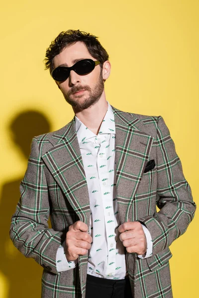 Stylish man in sunglasses touching checkered jacket on yellow background — Stock Photo