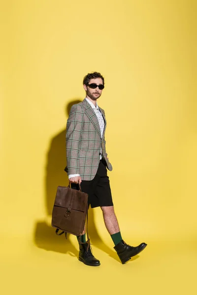 Trendy model in sunglasses and blazer holding backpack while walking on yellow background — Stock Photo