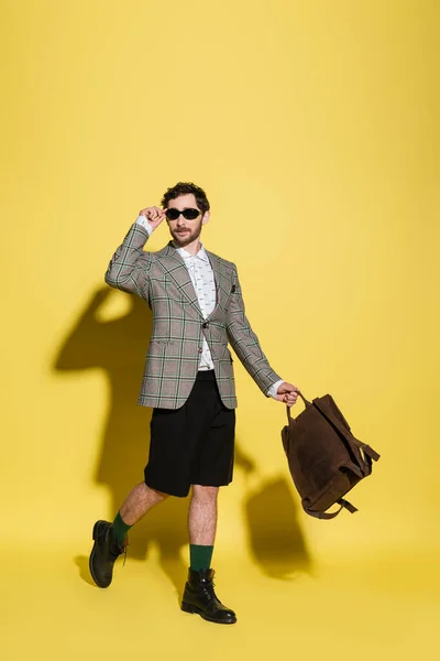 Fashionable model in sunglasses holding backpack and walking on yellow background — Stock Photo