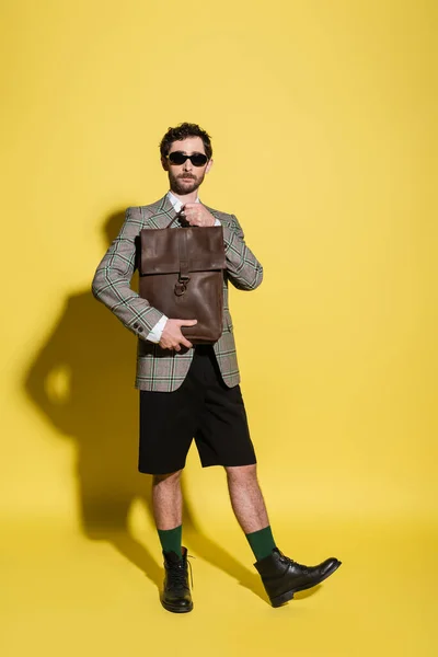 Full length of fashionable model in sunglasses and jacket holding brown bag on yellow background — Stock Photo