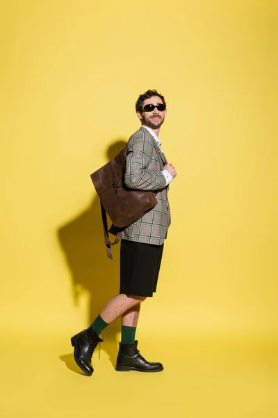 Улыбающийся и стильный мужчина в солнечных очках позирует с рюкзаком на желтом фоне — стоковое фото