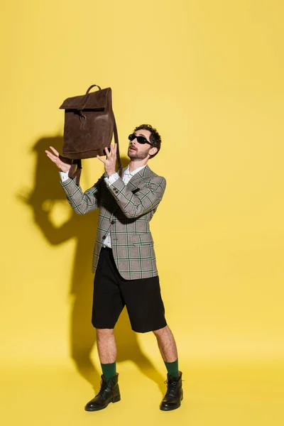 Повна довжина стильного чоловіка в сонцезахисних окулярах, що тримає коричневий рюкзак на жовтому фоні — стокове фото