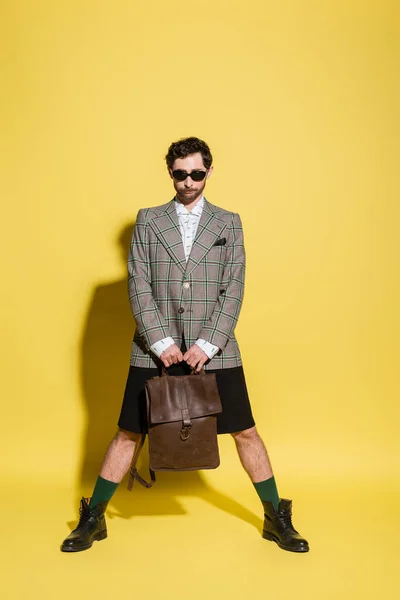 Fashionable model in jacket and sunglasses holding bag while standing on yellow background — Stock Photo