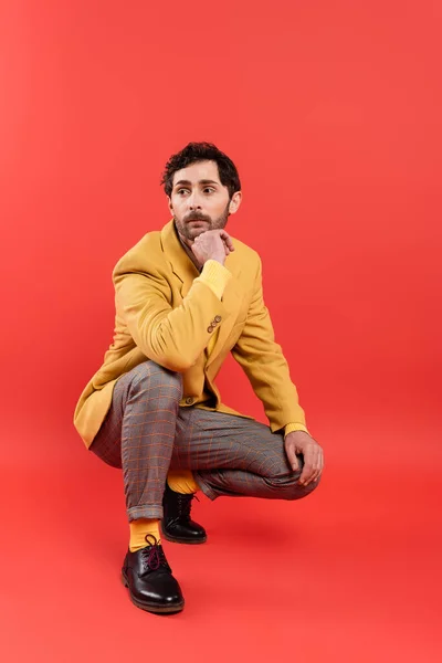 Homem barbudo na moda em calças xadrez e casaco amarelo posando no fundo vermelho coral — Fotografia de Stock