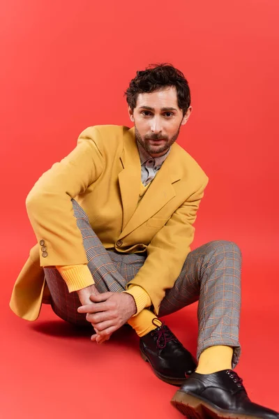 Hombre de moda y morena en chaqueta amarilla sentado sobre fondo rojo coral - foto de stock