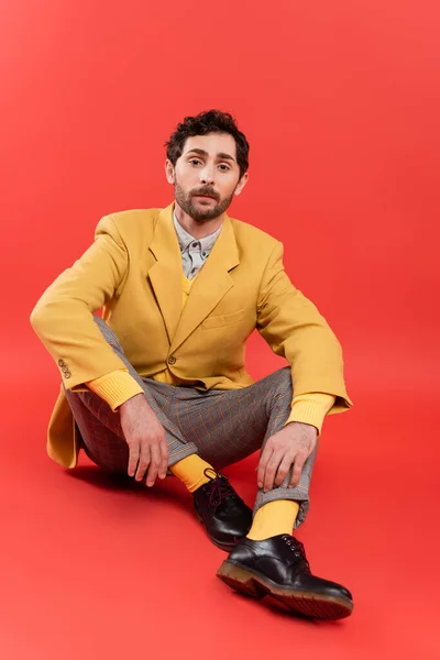 Modelo de moda en pantalones a cuadros y chaqueta amarilla sentado sobre fondo rojo - foto de stock