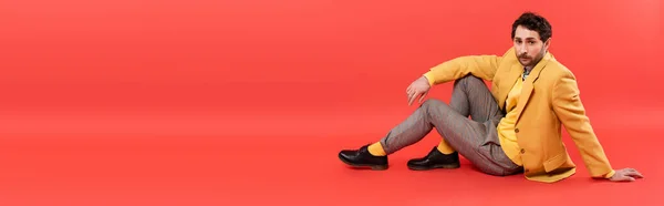 Homem morena na moda em casaco amarelo sentado no fundo vermelho coral, banner — Fotografia de Stock