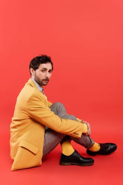 Elegante hombre morena en chaqueta amarilla y pantalones a cuadros sentados sobre fondo rojo coral - foto de stock