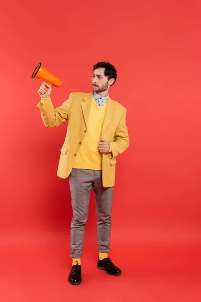 Stylish brunette man in jacket looking at loudspeaker on red background — Stock Photo