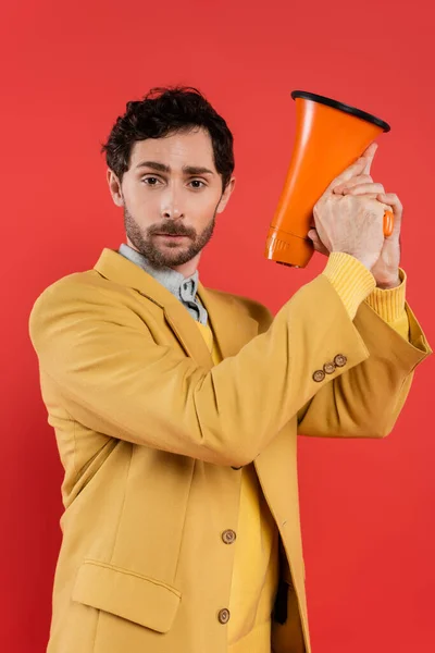 Portrait of fashionable man in jacket holding loudspeaker and looking at camera isolated on coral red — Stock Photo