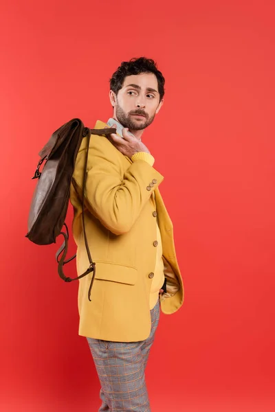 Модний чоловік в куртці тримає коричневий рюкзак ізольований на кораловому червоному — стокове фото