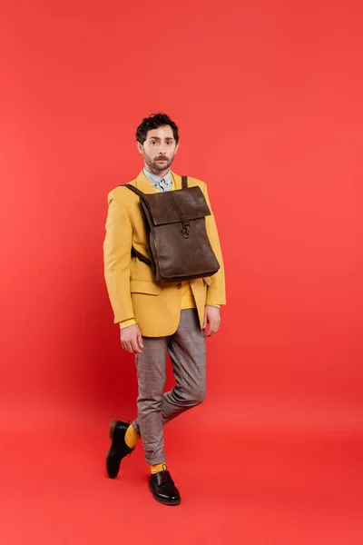 Volle Länge des modischen Modells mit braunem Rucksack über Jacke mit Blick auf die Kamera auf korallenrotem Hintergrund — Stockfoto