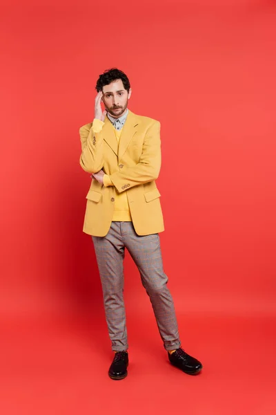 Longitud completa del hombre estresado en chaqueta amarilla y pantalones grises tocando la frente mientras está de pie sobre fondo de coral rojo - foto de stock