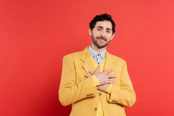 Thankful man in yellow blazer holding hands near chest while standing on red coral background — Stock Photo
