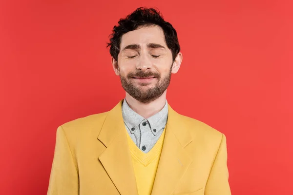 Pleased man in yellow blazer standing with closed eyes isolated on red coral background — Stock Photo