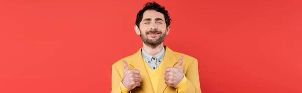 Pleased man in yellow blazer standing with closed eyes and showing thumbs up isolated on red coral, banner — Stock Photo