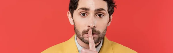 Homme en blazer jaune tenant doigt près des lèvres tout en montrant un signe de silence isolé sur fond de corail rouge, bannière — Photo de stock