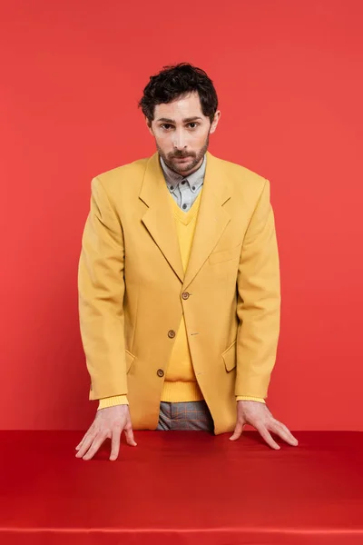 Uomo in elegante giacca gialla in piedi vicino alla scrivania rossa e guardando la fotocamera su sfondo corallo — Foto stock