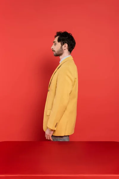 Seitenansicht eines bärtigen Mannes in stylischem gelben Blazer, der neben rotem Schreibtisch auf korallenfarbenem Hintergrund steht — Stockfoto