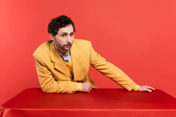 Man in stylish yellow blazer leaning on table and looking at camera on red coral background — Stock Photo