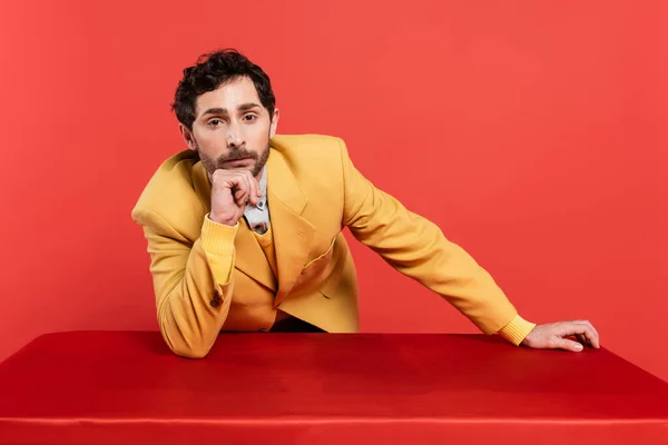 Homem pensativo no elegante blazer amarelo inclinado na mesa e olhando para a câmera isolada no fundo de coral vermelho — Fotografia de Stock