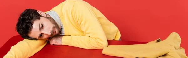 Tipo cansado en amarillo jersey de manga larga acostado en escritorio rojo cerca de chaqueta aislado en el fondo de coral, pancarta - foto de stock