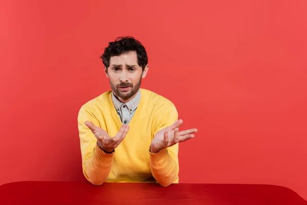 Confused man in yellow long sleeve jumper sitting at table and gesturing on coral background — Stock Photo