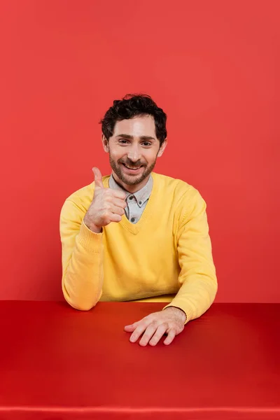Счастливый мужчина в желтых джинсах с длинными рукавами, показывающий большой палец вверх на красном коралловом фоне — стоковое фото