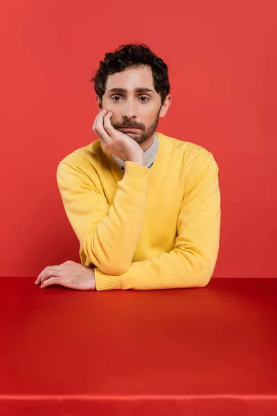 Frustrated man in yellow long sleeve jumper sitting at desk on red coral background — Stock Photo