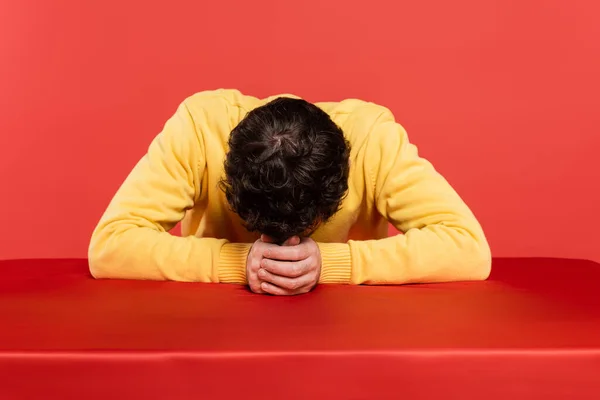 Homem encaracolado em amarelo jumper manga longa sentado com cabeça curvada na mesa isolada no fundo de coral — Fotografia de Stock