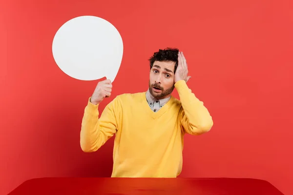 Worried man in yellow long sleeve jumper holding blank speech bubble on red coral background — Stock Photo