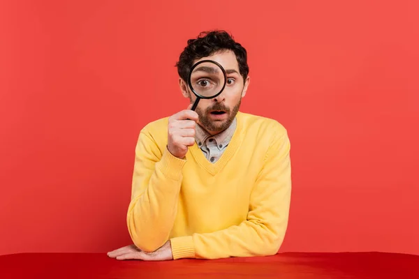 Scrupulous man in yellow long sleeve jumper holding magnifying glass on red coral background — Stock Photo
