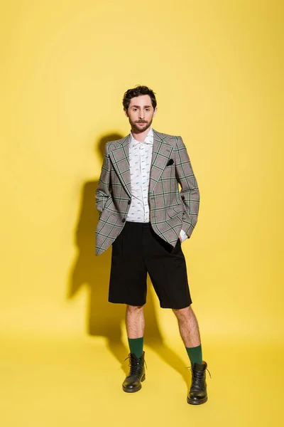 Full length of charming guy in grey checkered blazer and shorts standing with hands in pockets on yellow background — Stock Photo