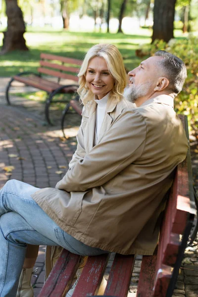 Mujer rubia positiva sentada cerca de marido maduro en el banco en el parque - foto de stock