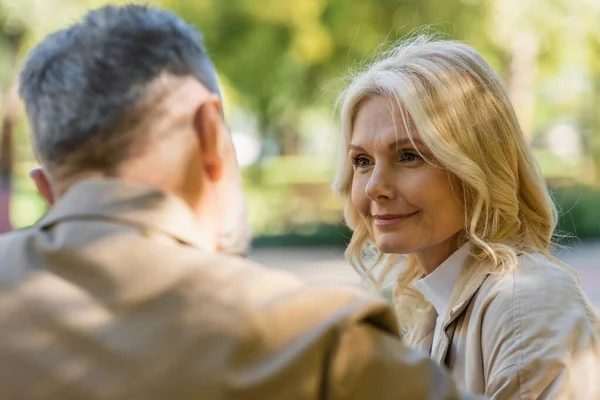 Smiling blonde woman looking at blurred husband in spring park — Stock Photo