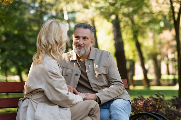 Sonriente hombre maduro cogido de la mano de la esposa rubia mientras está sentado en el banco en el parque - foto de stock