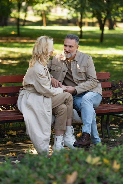 Mature couple in spring outfit holding hands while sitting on bench in park — Stock Photo