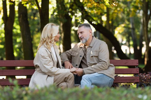 Mature man talking and holding hand of blonde wife while sitting on bench in park — Stock Photo