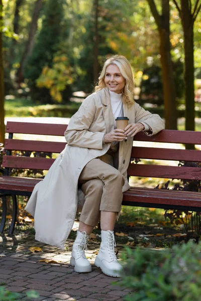 Сверхрадостная блондинка в плаще держит кофе, чтобы пойти, сидя на скамейке в весеннем парке — стоковое фото
