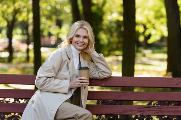 Cheerful mature woman in trench coat holding coffee to go and sitting on bench in park — Stock Photo