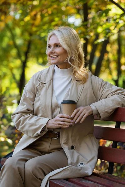 Беззаботная блондинка держит бумажный стаканчик, сидя на скамейке в весеннем парке — стоковое фото